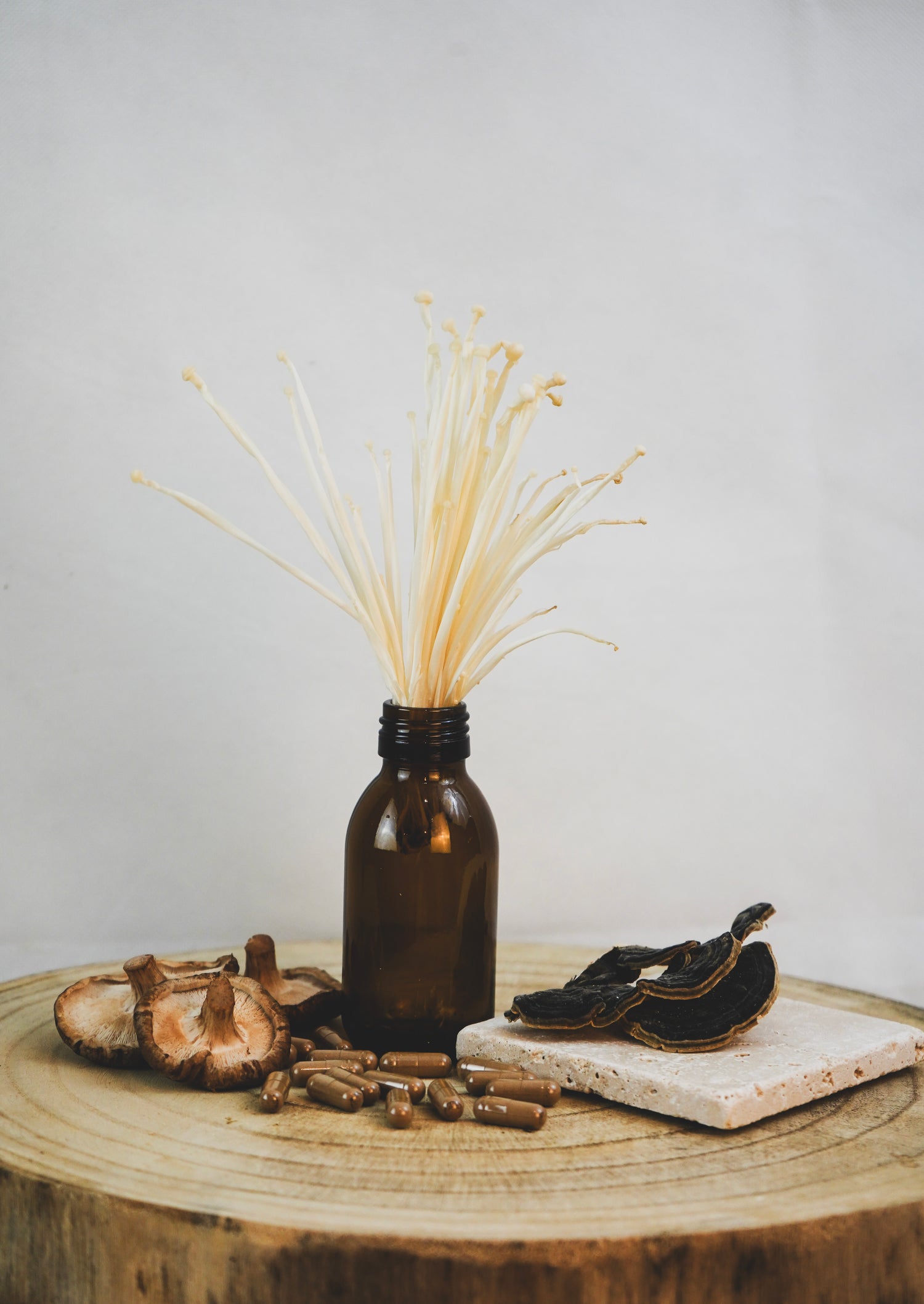Braunes Glasfläschchen mit Enoki-Pilzen, daneben Shiitake-Pilze, Vitalpilz-Kapseln und getrocknete Pilzscheiben auf Holz, natürliche Produktpräsentation